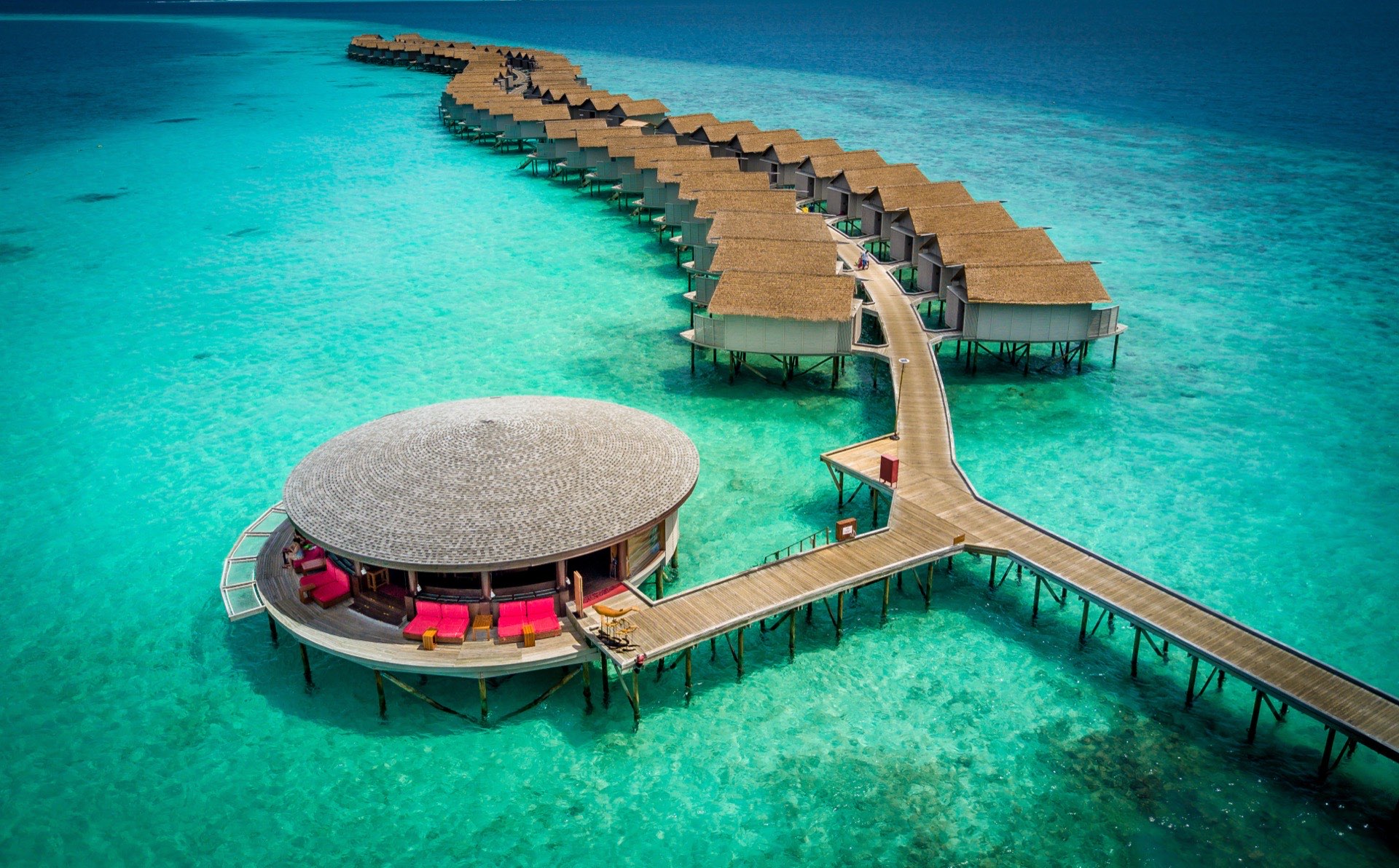 Aerial view of Centara Ras Fushi: a small palm-covered island ringed by white sand, with rows of thatched overwater villas reaching out across a turquoise lagoon.